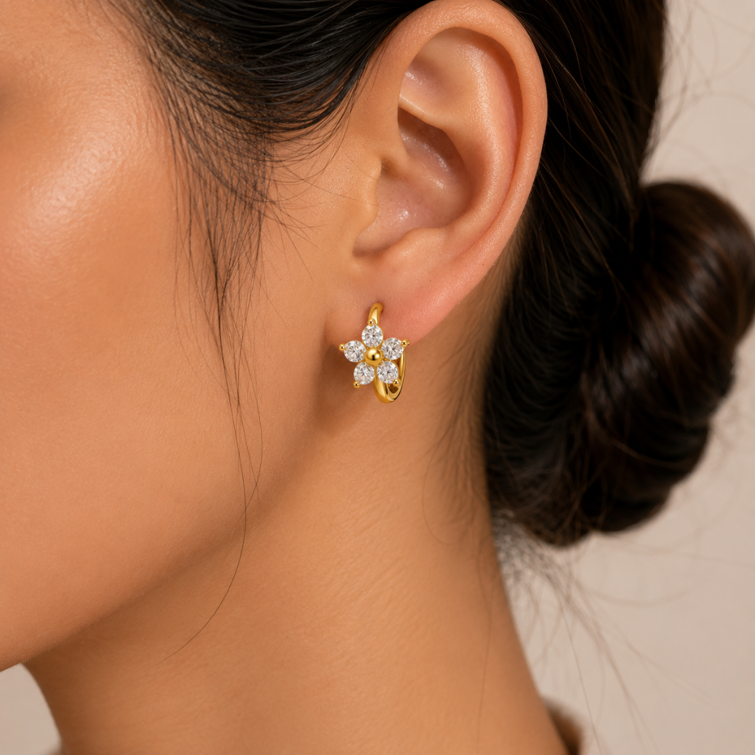 A close-up of a woman's ear wearing a small gold flower-shaped stud earring with clear gemstones, against a soft beige background.