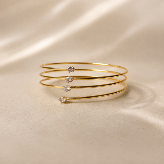 Three thin gold bangle bracelets stacked together, each decorated with a small clear round gemstone, displayed on a soft beige satin fabric background.