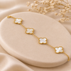 A delicate gold chain bracelet with four white mother-of-pearl four-leaf clover motifs, displayed on a beige oval jewelry stand with soft satin fabric and dried flowers in the background.
