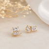 A pair of gold stud earrings featuring a stacked design with a square princess-cut clear gemstone on top of a marquise-cut clear gemstone, displayed on a light beige textured surface with dried baby's breath flowers.