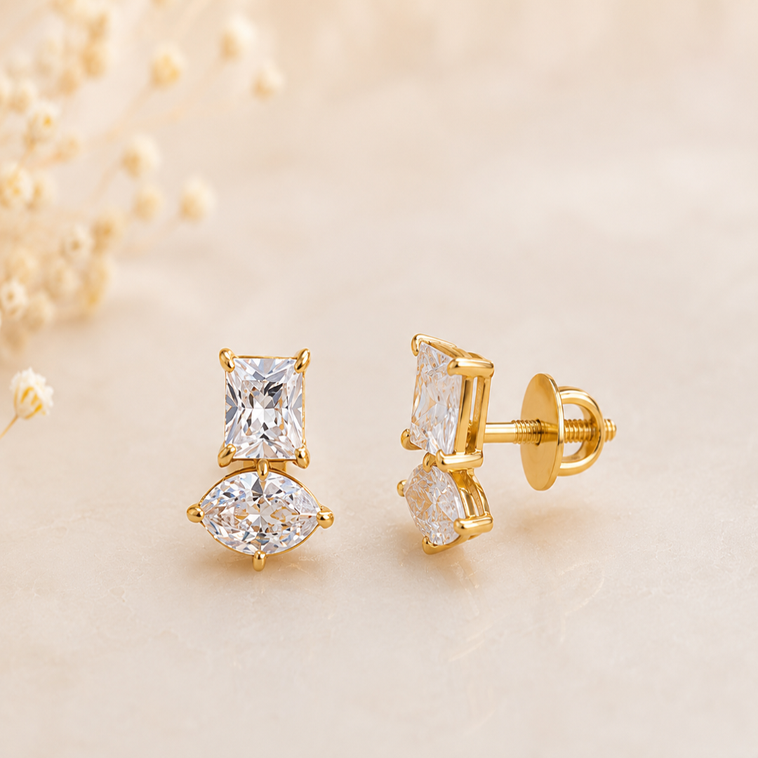 A pair of gold stud earrings featuring a princess-cut clear gemstone and a marquise-cut clear gemstone, displayed on a soft beige surface with blurred dried flowers in the background.