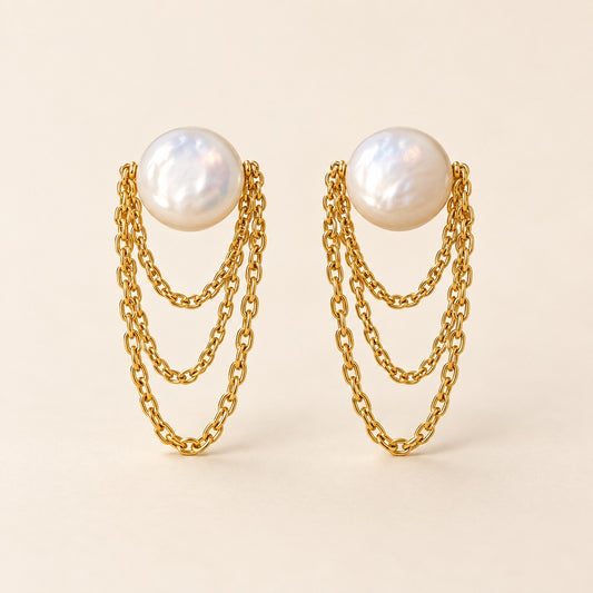 A pair of gold-toned dangle earrings featuring a large white freshwater pearl at the top of each earring, with three cascading chain layers below, set against a solid light beige background.