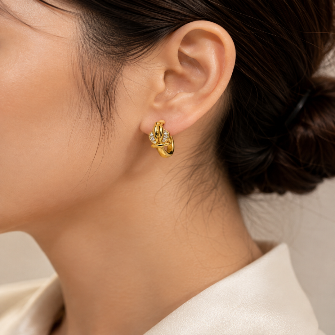 A close-up of a model's ear wearing a small gold huggie hoop earring with clear gemstones, set against a soft beige background.