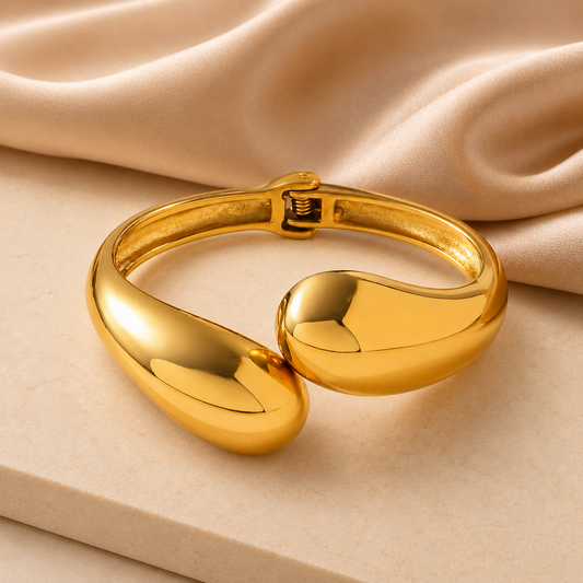 A polished gold-toned metal cuff bracelet with a smooth, rounded organic shape, displayed on a beige surface with draped satin fabric in the background.