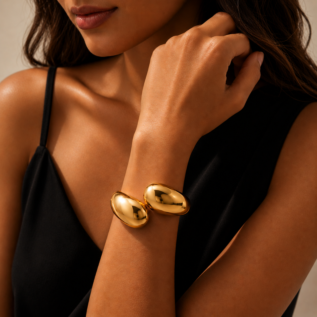 A close-up lifestyle shot of a model wearing a chunky, polished gold cuff bracelet with two connected oval links. The model is wearing a black camisole, and the background is a soft, warm neutral beige.
