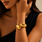 A close-up of a woman wearing a polished gold cuff bracelet on her wrist, dressed in a black sleeveless top against a warm beige background.