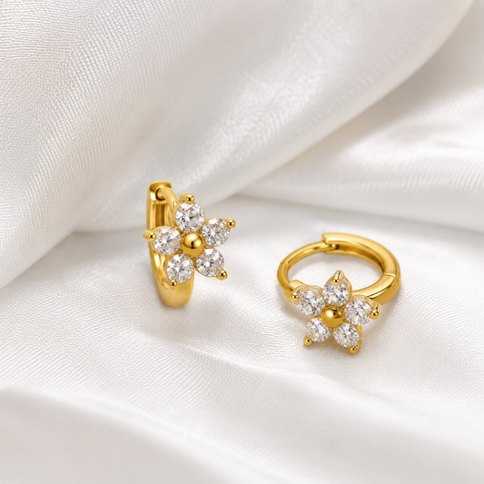 A pair of gold huggie hoop earrings with a flower design set with clear round gemstones, displayed on a draped white satin fabric.