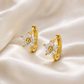 A pair of gold huggie hoop earrings with a flower-shaped cluster of clear stones and a central gold bead, displayed on a draped cream satin fabric.