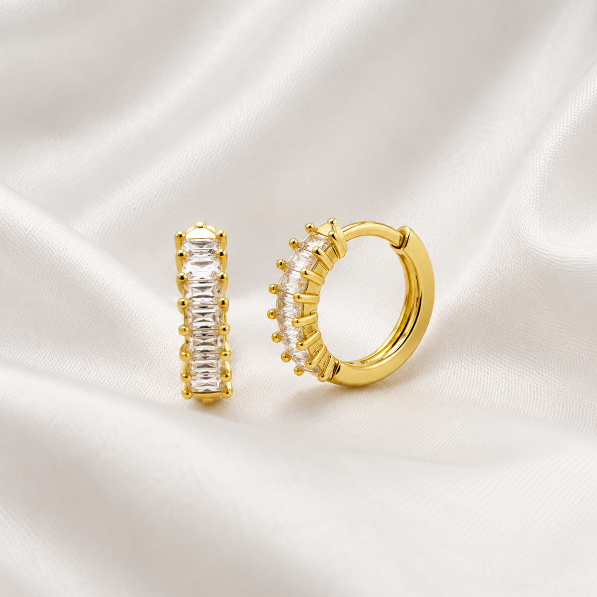 A pair of gold hoop earrings displayed on a draped white satin fabric. One earring is shown vertically with clear baguette-cut stones, and the other is shown horizontally with clear stones set in a scalloped border.