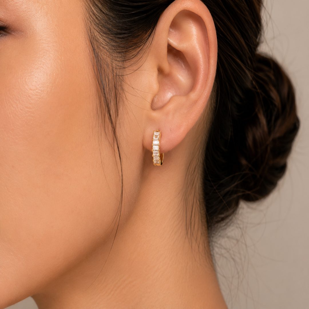 A close-up of a model's ear wearing a small gold huggie hoop earring adorned with clear rectangular baguette crystals, set against a neutral beige background.