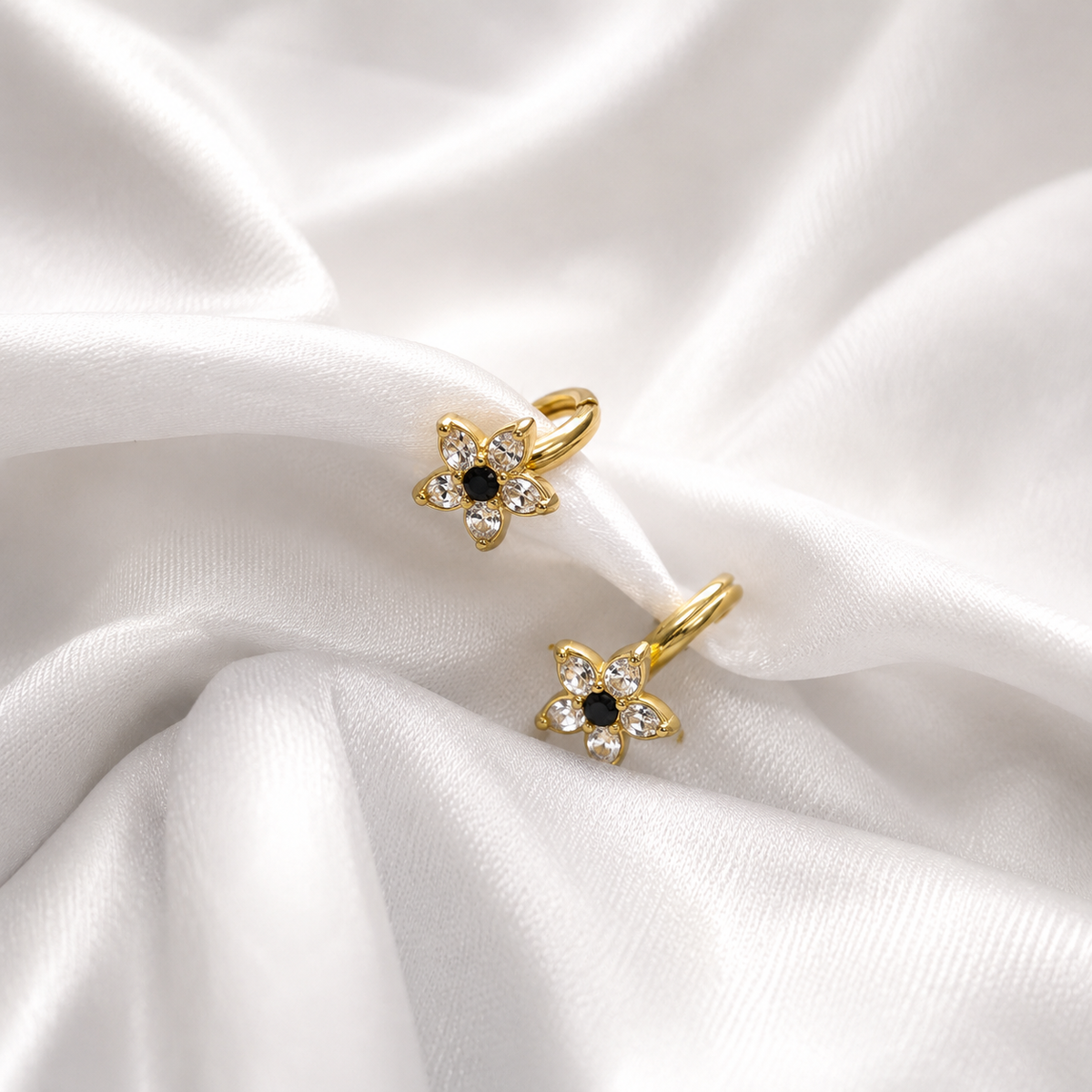 A pair of gold flower-shaped hoop earrings with clear rhinestones and a small black center stone, displayed on draped white satin fabric.