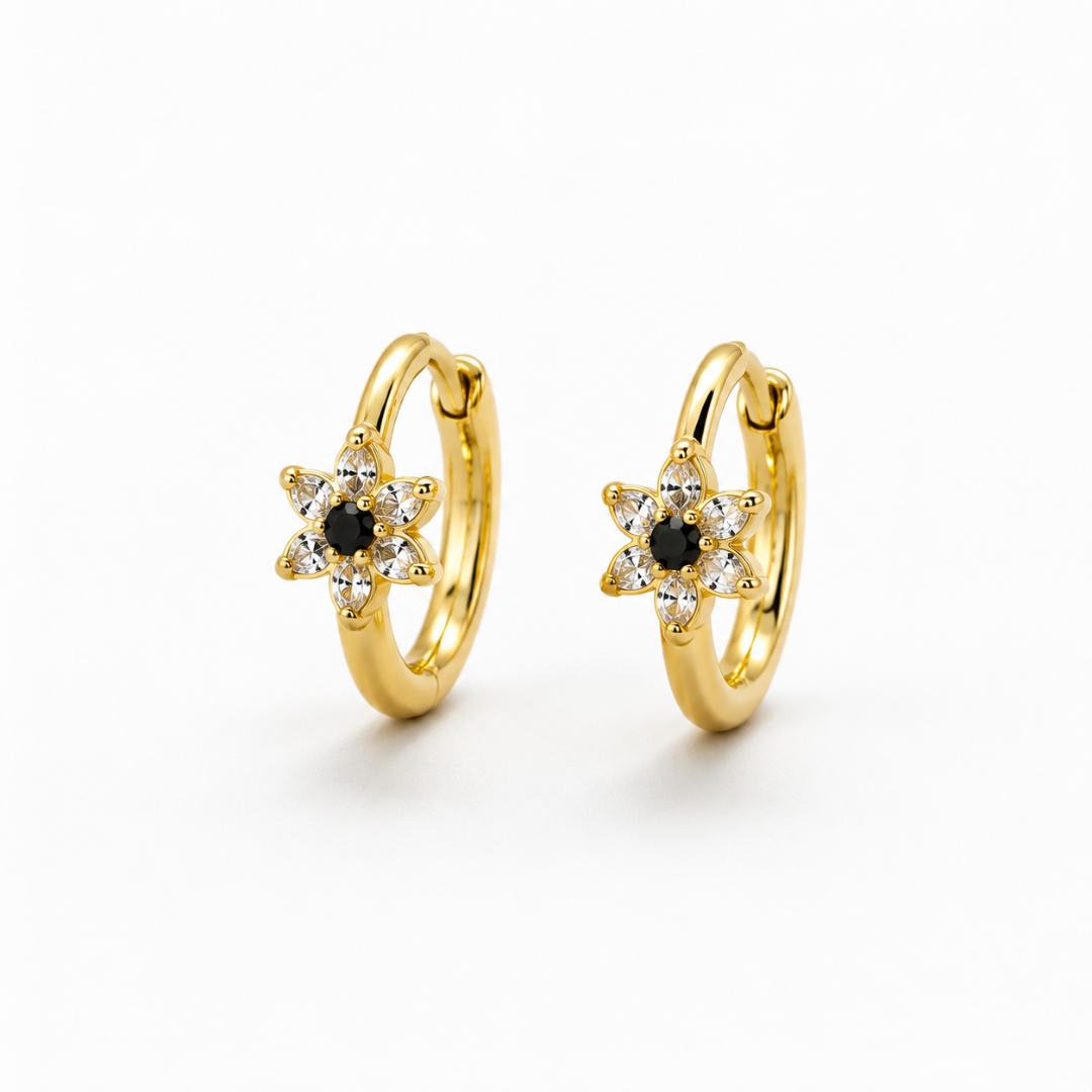 A pair of gold huggie hoop earrings with a star-shaped floral center, featuring a small black round stone surrounded by clear stones, set against a plain white background.