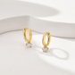 A pair of gold huggie hoop earrings with a clear round-cut gemstone dangling from the bottom of each hoop, displayed on a clean white surface with soft shadows.