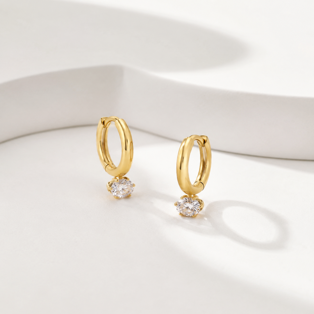 A pair of gold huggie hoop earrings with a clear round-cut gemstone dangling from the bottom of each hoop, displayed on a clean white surface with soft shadows.