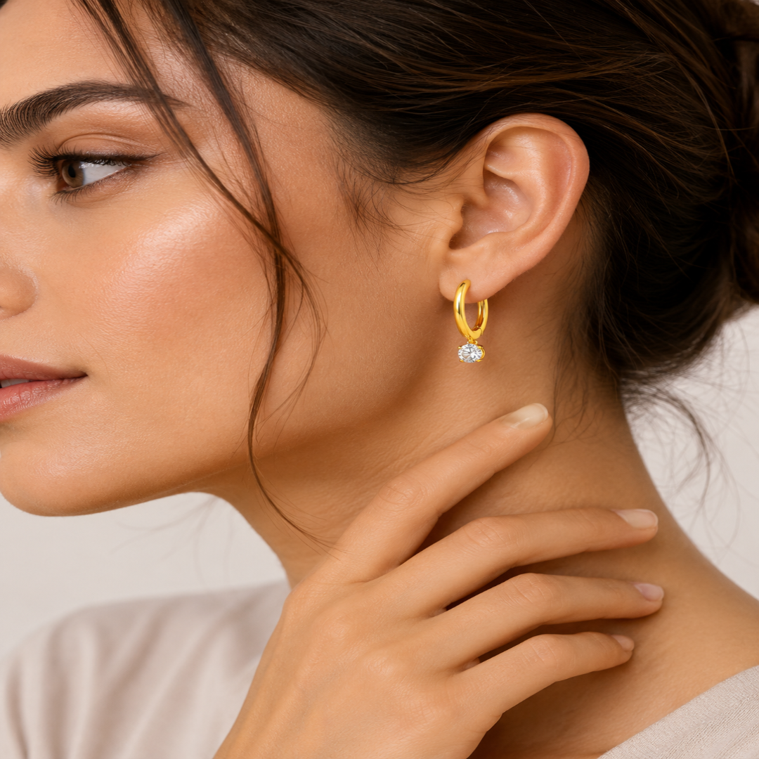 A close-up lifestyle shot of a woman wearing a delicate gold huggie hoop earring with a small clear gemstone dangling at the bottom. The image highlights the earring on her ear, with soft, warm lighting and a clean, neutral background.