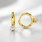A pair of gold huggie hoop earrings, one standing vertically and the other lying horizontally, each featuring a round brilliant-cut clear gemstone in a prong setting, displayed on a clean white surface with soft shadows.