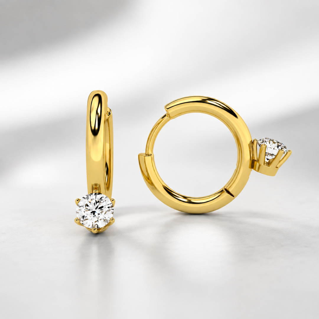 A pair of gold huggie hoop earrings, one standing vertically and the other lying horizontally, each featuring a round brilliant-cut clear gemstone in a prong setting, displayed on a clean white surface with soft shadows.