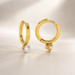 A pair of gold huggie hoop earrings with a small clear gemstone in a prong setting, displayed on a neutral beige background.