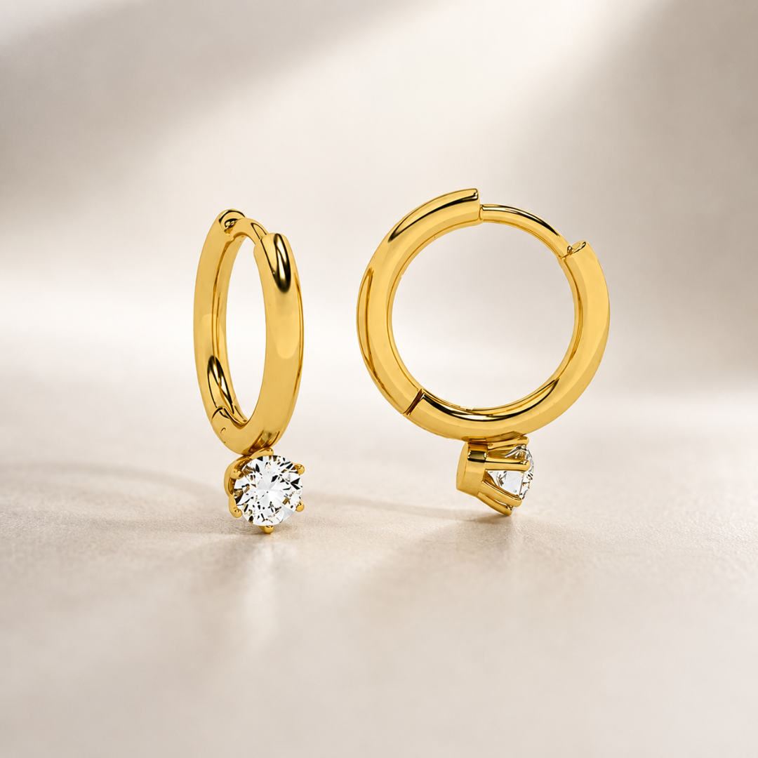 A pair of gold huggie hoop earrings with a small clear gemstone in a prong setting, displayed on a neutral beige background.
